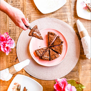 a hand serving Carolina's brownie on a pink plate. In the background a wooden table set up with pink placemats and white napkins and pink flowers