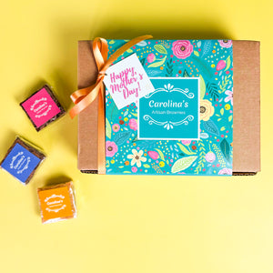 Top view of a craft box with teal sleeve with a flower pattern and Happy Mother's Day tag and ribbon. On the left side, three scattered brownies with colourful labels.