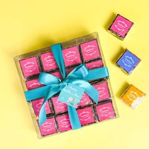 Square box showing  sixteen brownies with pink labels and teal ribbon and Happy Mother's Day tag. On the left side, three brownies with colourful labels scattered.